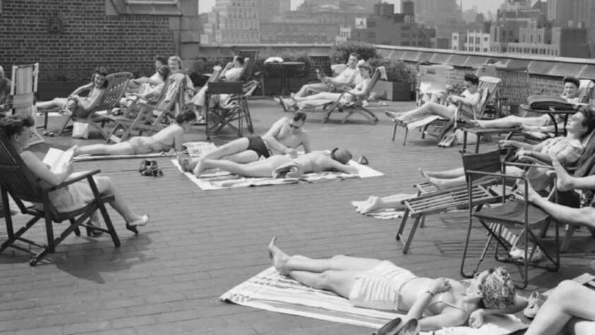 Black and white photo of people sunbathing on a city rooftop, lying on towels or sitting in lounge chairs, with tall buildings visible in the background.