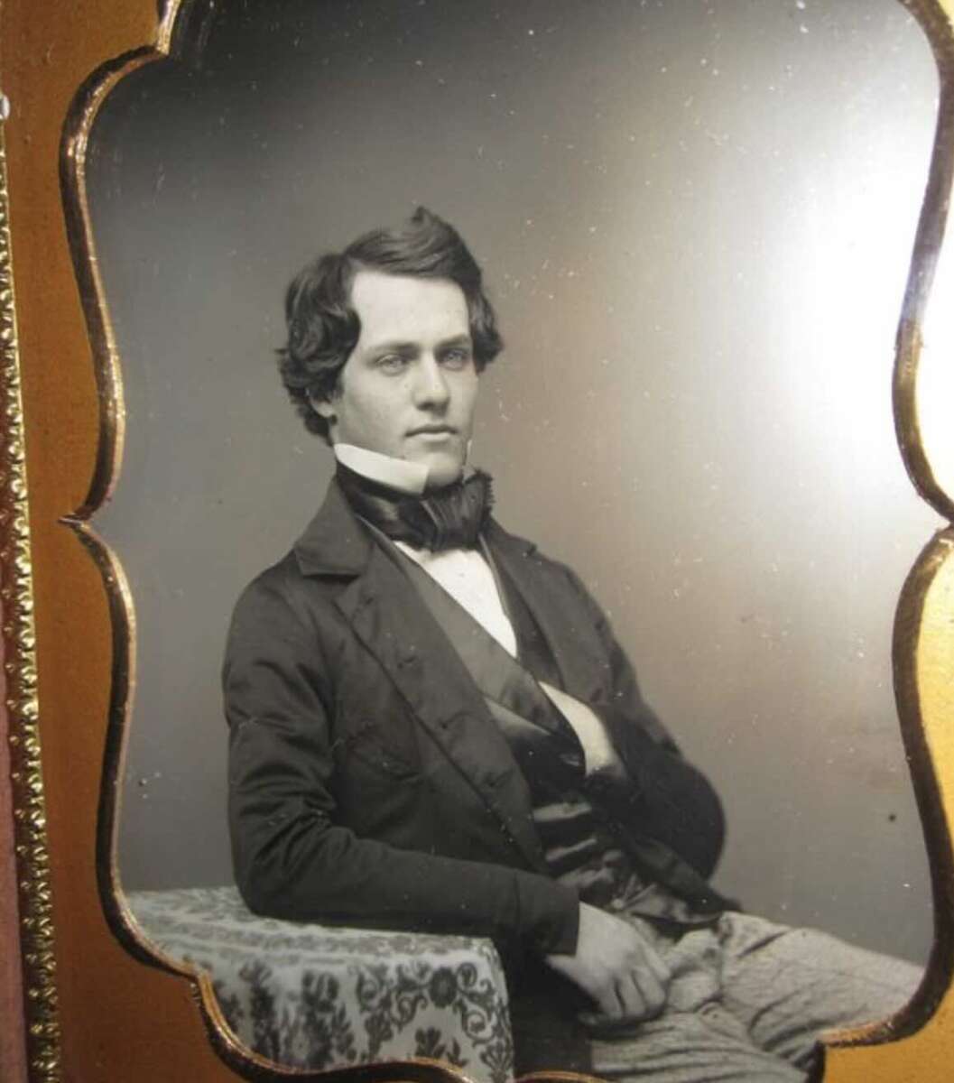 A young man with wavy dark hair sits in a formal pose, wearing a dark jacket, waistcoat, and bow tie. He rests one arm on a patterned table and the other hand inside his jacket, against his chest.