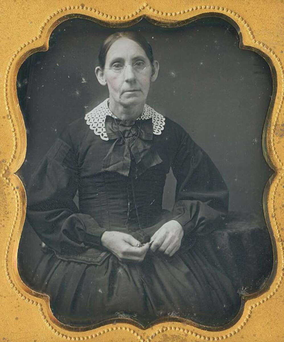 A seated woman in a dark, long-sleeved dress with a lace collar and a bow at her neck poses for a formal portrait against a dark background. The photo is framed in an ornate, gold-edged case.