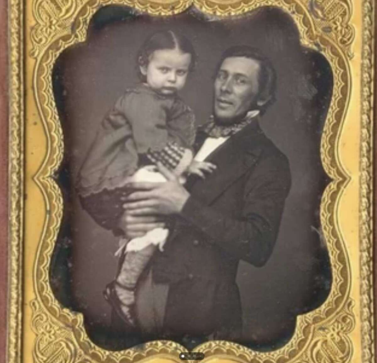 A vintage sepia photograph in an ornate gold frame shows a man in a suit holding a young child in a dress. The man gazes slightly off-camera while the child looks directly at the lens, both with neutral expressions.