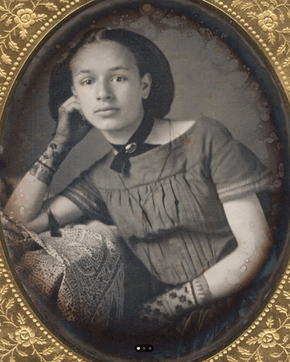 A vintage, sepia-toned portrait of a young woman posing with her head resting on her hand. She wears lace gloves, a dress with pleats, and a choker. The photo is framed in an ornate, gold oval frame.