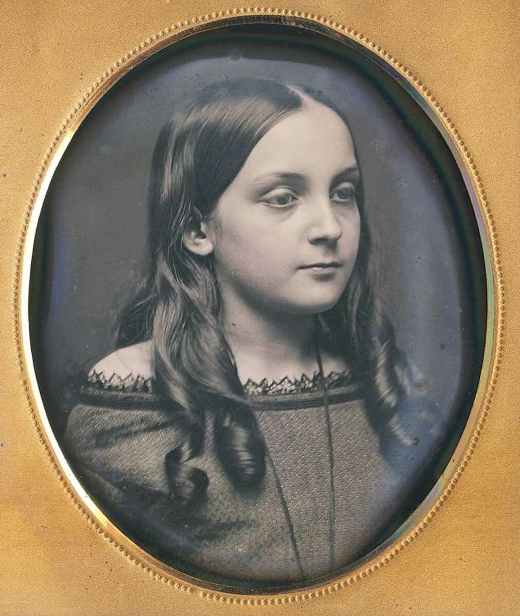 A vintage black-and-white portrait of a young girl with long, wavy hair, wearing an off-shoulder dress, framed in an oval, gold-colored frame. She gazes slightly to the side with a calm expression.