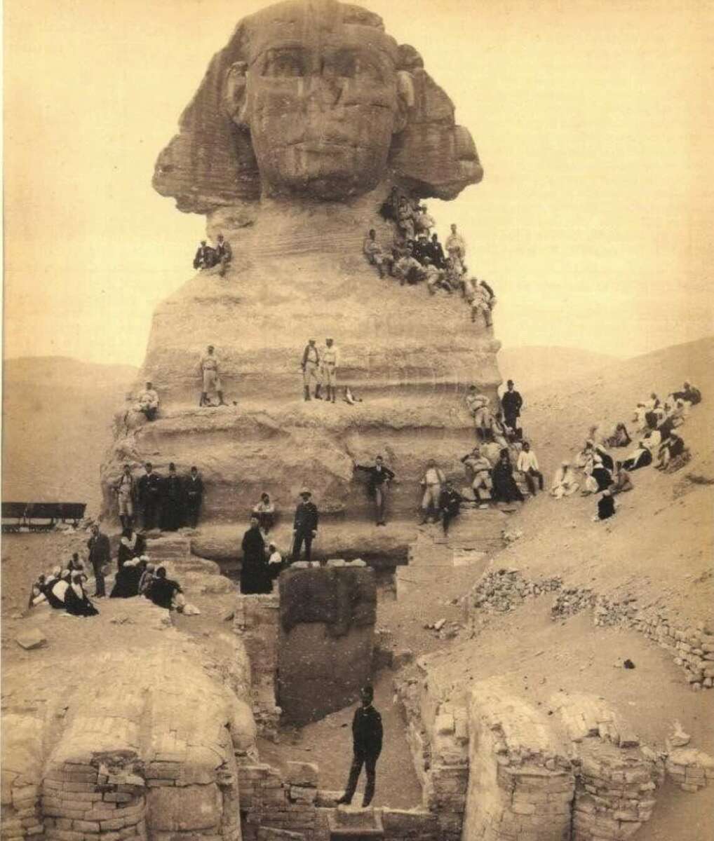 A sepia-toned historical photo shows dozens of people posing on and around the partially excavated Great Sphinx of Giza, with sand and ruins in the background.
