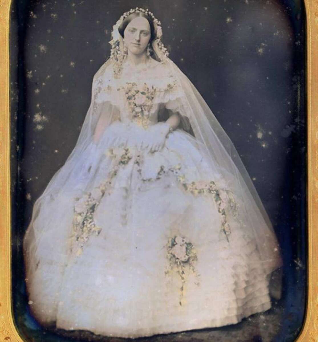 A sepia-toned vintage photograph of a woman in a voluminous white wedding dress with floral details and a long veil, posing formally and looking at the camera. The image has a soft, ethereal quality.