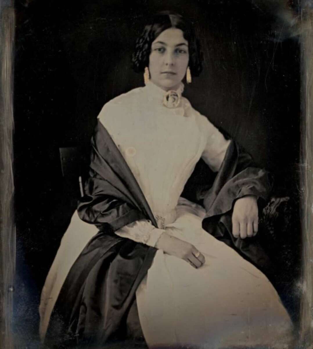 A seated woman in a long white dress with dark shawl, wearing earrings and a brooch, gazes calmly at the camera in this vintage, color-tinted portrait photograph.