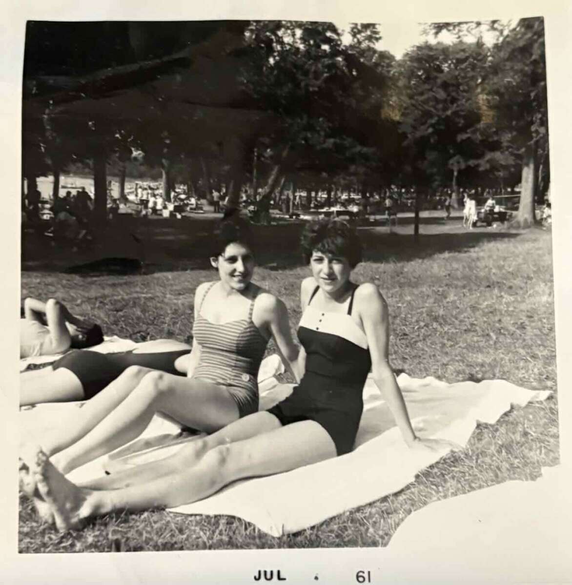 Two women in vintage swimsuits sit on towels on grass in a park, smiling at the camera. Other people relax in the background among trees. The photo is black and white, dated July 1961.