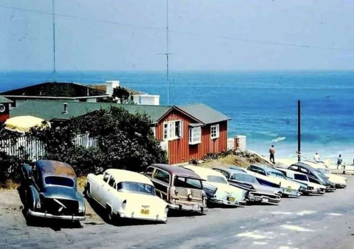 A row of vintage cars is parked along a seaside road, with a red wooden building and the ocean in the background on a sunny day. A few people can be seen near the water’s edge.
