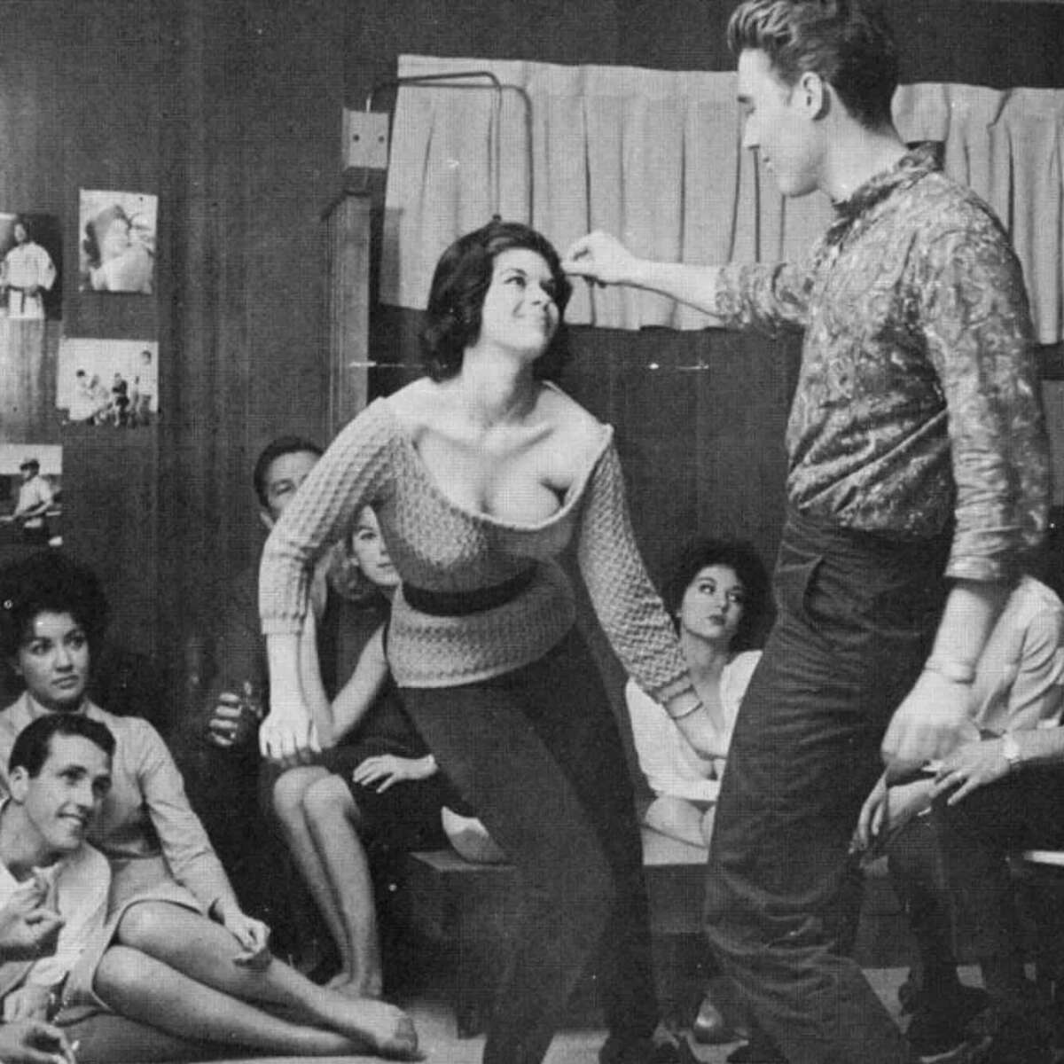 A group of young adults sit and watch as a woman and man dance together in the center of a room, smiling and enjoying themselves. The setting appears to be a casual indoor gathering or party.