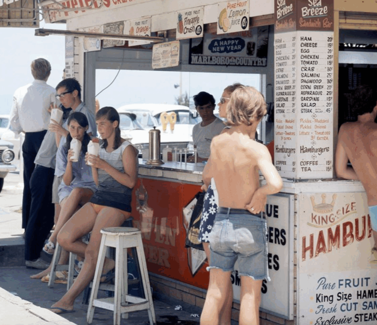 A group of young people in swimwear gather at a colorful beachside snack stand, drinking milkshakes and talking. Vintage cars and a sunny beach can be seen in the background.