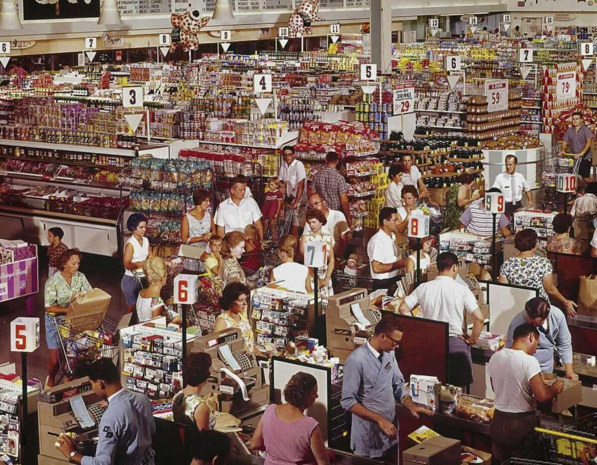 A busy supermarket scene shows crowded checkout lanes with numbered signs, cashiers scanning items, shoppers waiting with full carts, and shelves stocked with various groceries. The store is bustling with activity.
