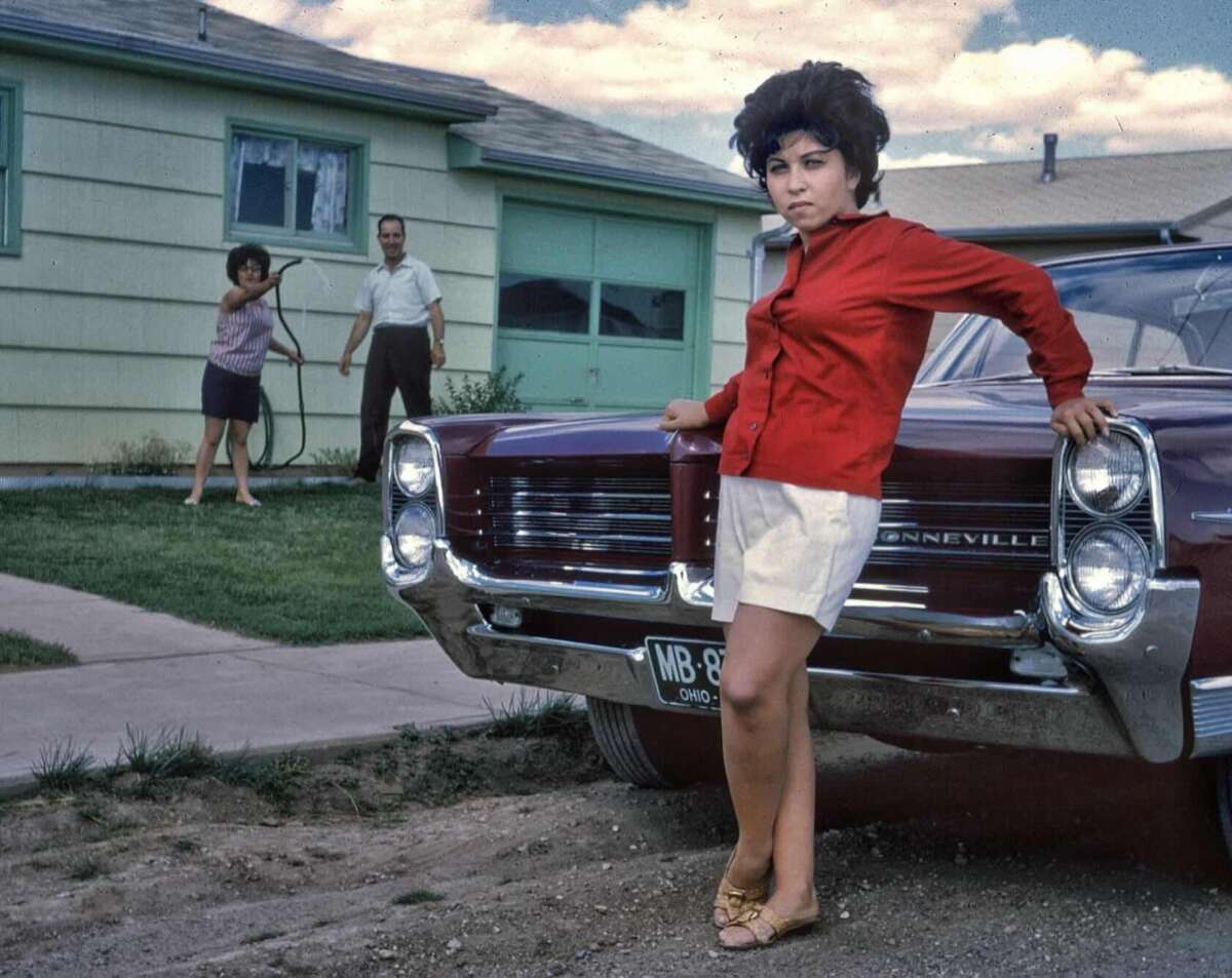 A woman in a red shirt and white shorts leans against a maroon Pontiac Bonneville parked in front of a house, while a man and woman stand in the yard, the woman holding a garden hose.