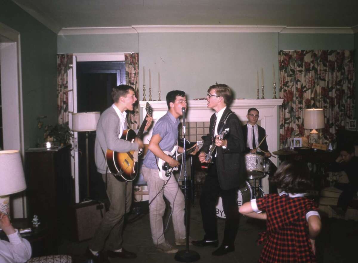 Four young men play instruments and sing into a microphone in a living room, with a child in a red plaid dress dancing in the foreground. Curtains and lamps are visible, creating a cozy, informal atmosphere.