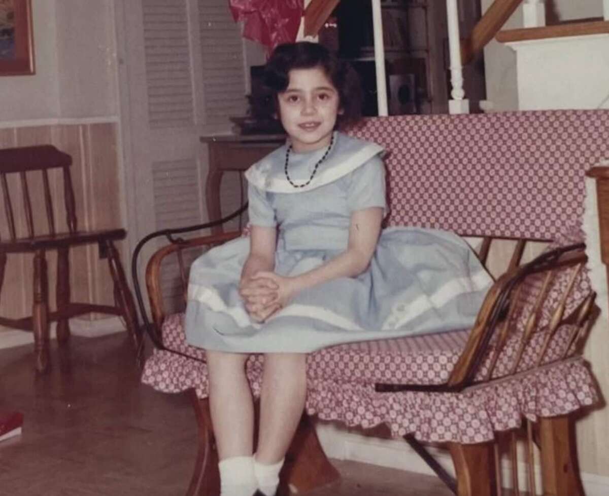 A young girl in a blue dress with a white collar and necklace sits on a patterned bench indoors, hands clasped in her lap, smiling at the camera. The setting appears to be a cozy, well-lit home.