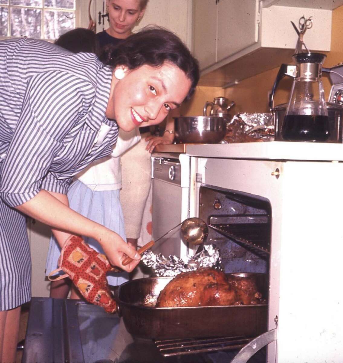 A woman in a striped dress and oven mitt smiles while basting a roast in the oven. Two others, one woman and a child, stand behind her in a kitchen with yellow walls and white cabinets.