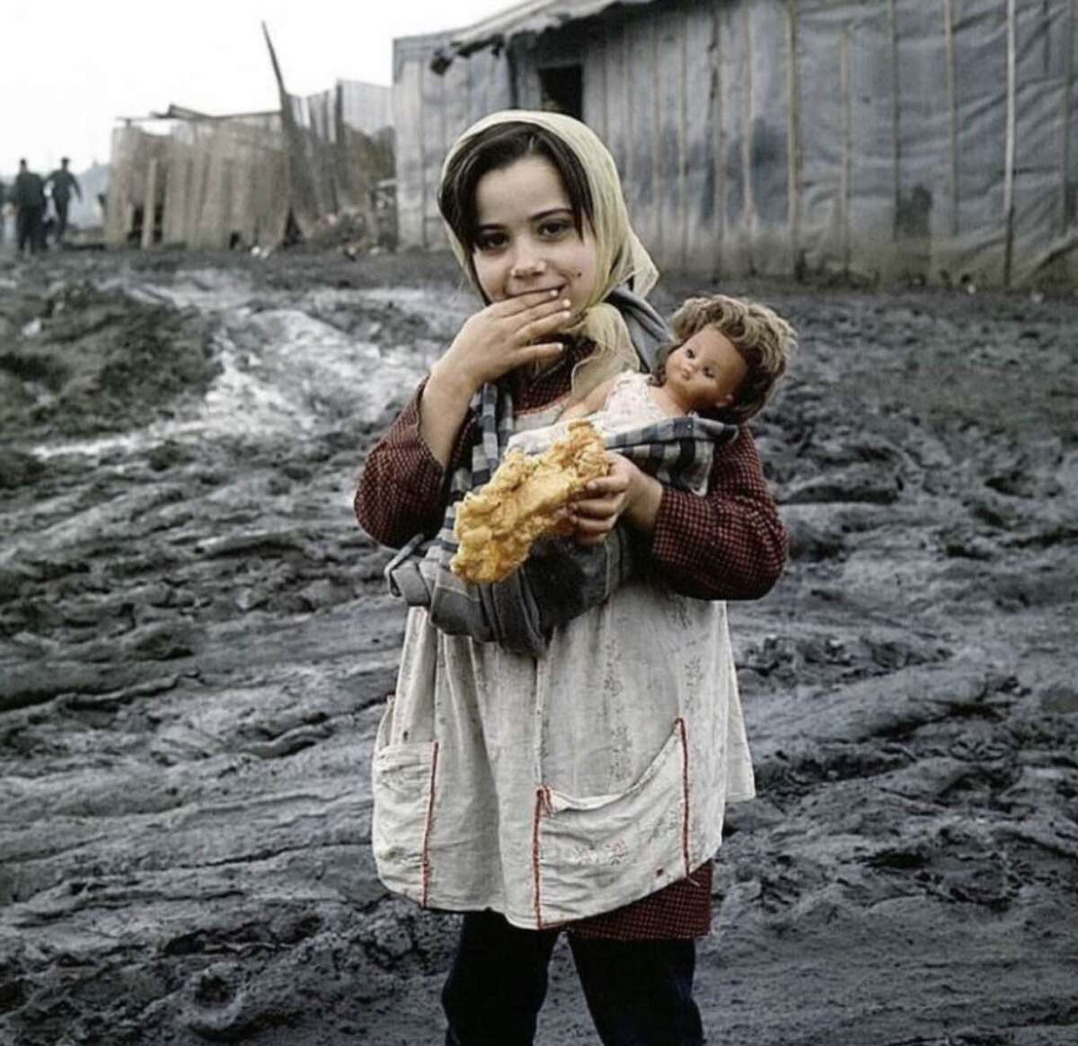 A young girl stands in a muddy area, holding bread in one hand and a doll in the other. She wears a headscarf and apron, with tents and blurred figures visible in the background.