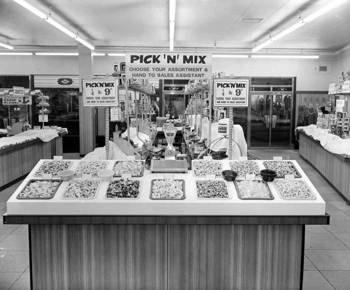 Black and white photo of a store’s Pick ‘N’ Mix candy counter with various sweets in trays. Signs instruct customers to choose their assortment and hand it to a sales assistant. The store interior and shoppers are visible in the background.