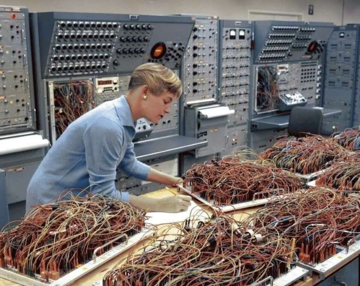 A woman in a blue blouse works with large panels of tangled wires in a 1960s-era computer lab, surrounded by vintage control panels filled with switches and dials.