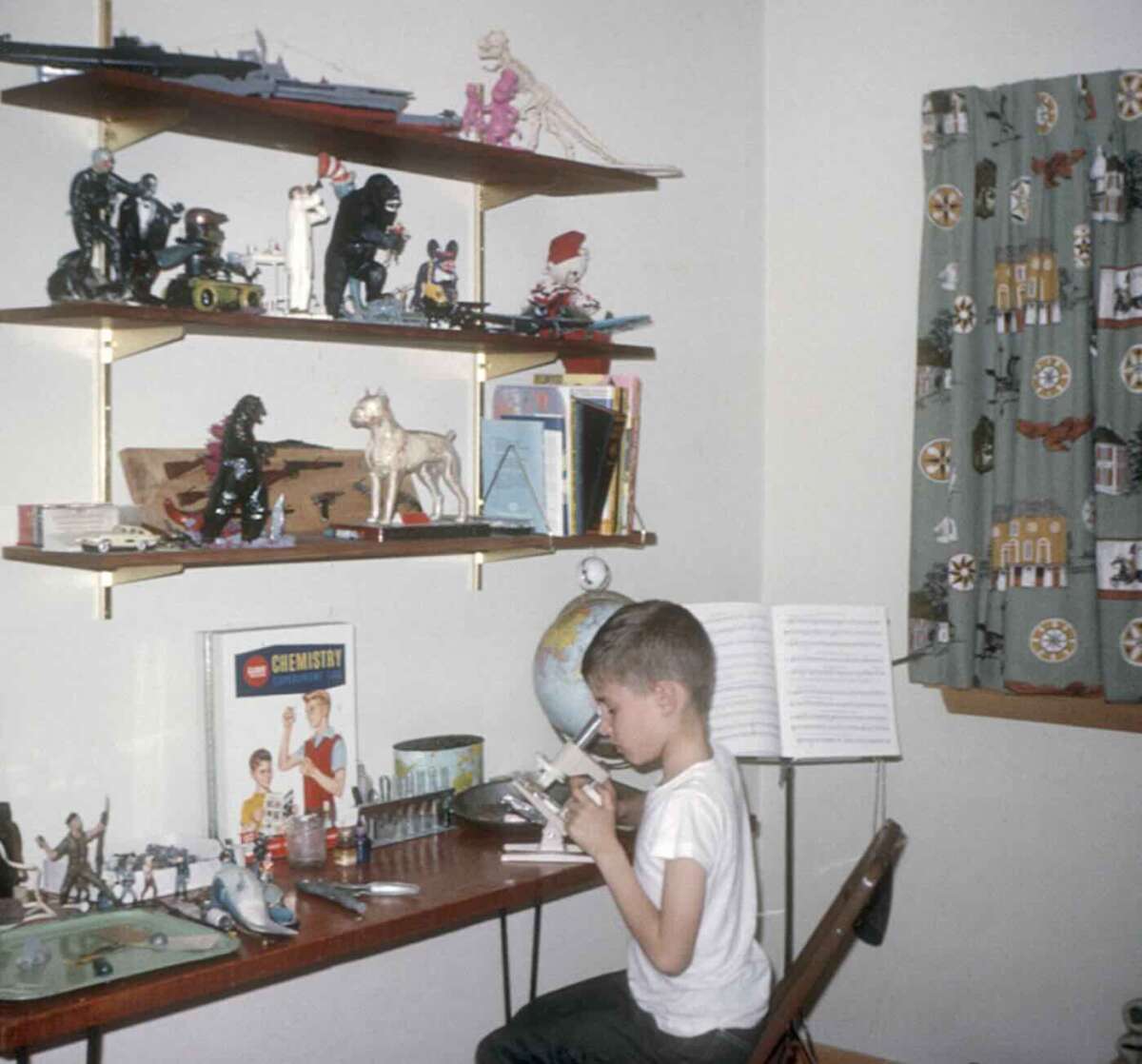 A young boy sits at a desk in a bedroom, building a model airplane. Shelves above his desk hold toy figures, model ships, and animals. To his left is a globe and a chemistry set; a window with patterned curtains is nearby.