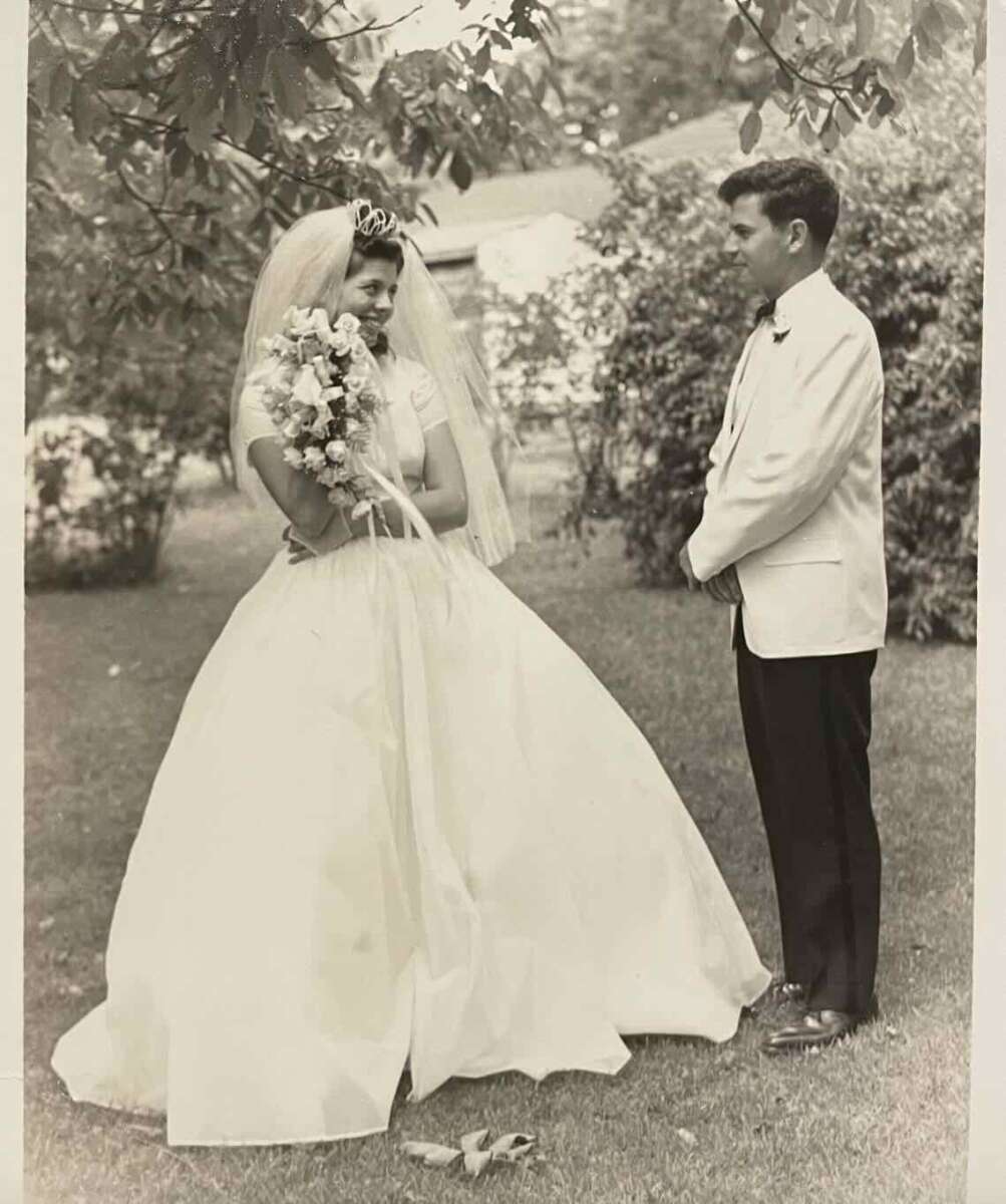 A bride in a white gown and veil holding a bouquet stands outdoors, smiling at a man in a white tuxedo jacket and dark pants. They appear to be at a wedding, standing on grass with trees and houses in the background.
