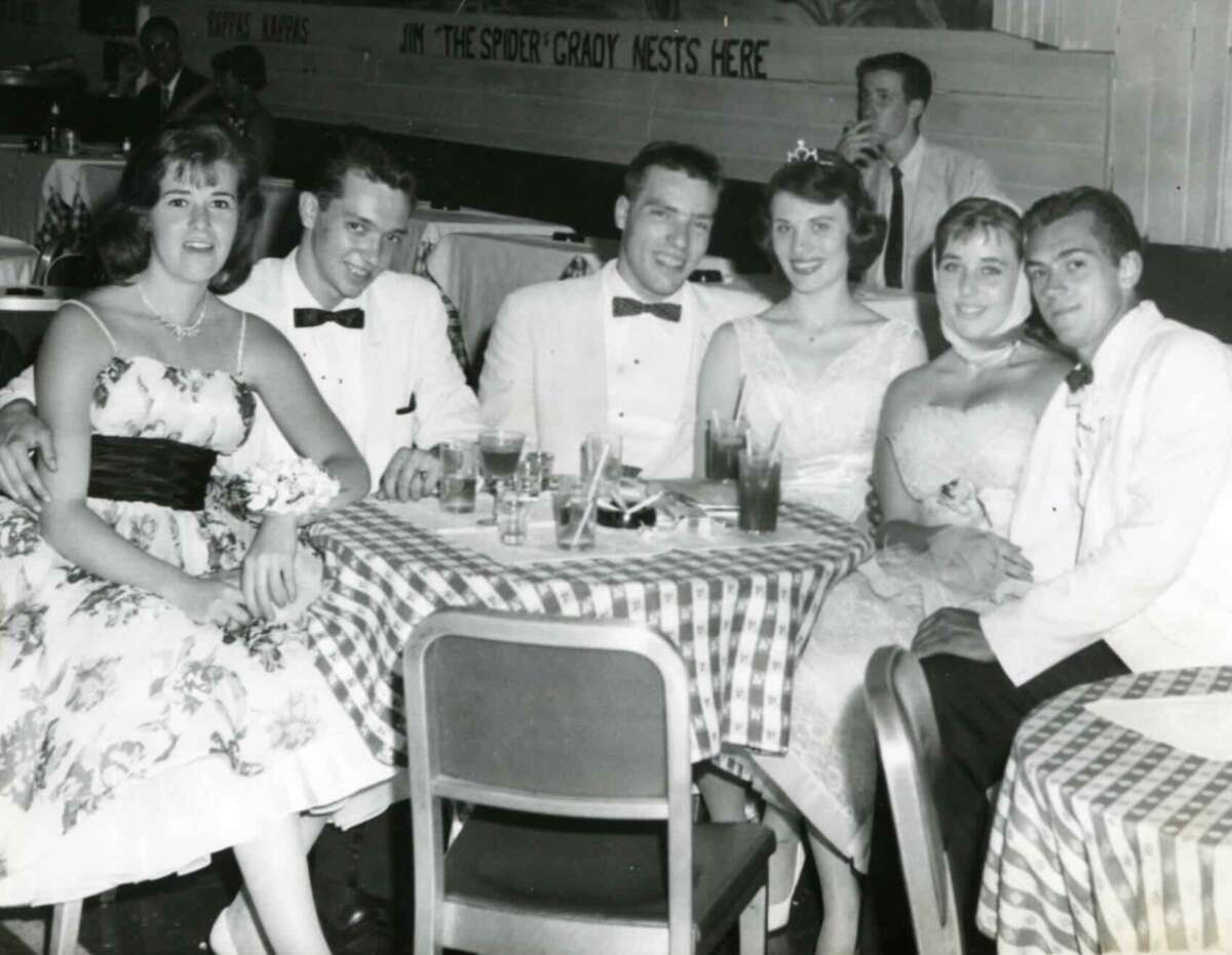 Six young adults dressed formally sit around a table with drinks at a checkered tablecloth venue, smiling at the camera. A sign on the wall behind them reads "THE SPORE'CROW NESTS HERE.