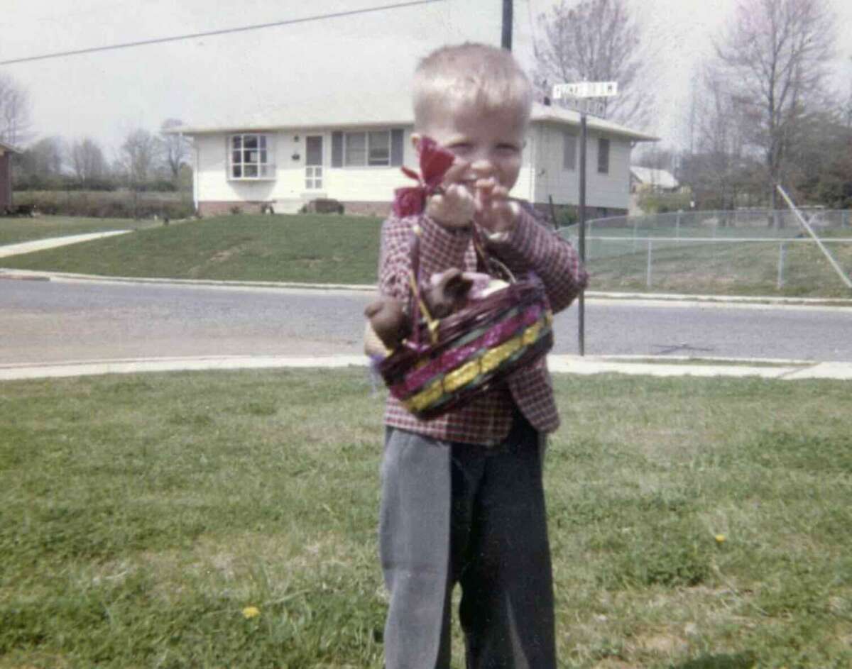 A young boy stands on a grassy lawn holding a colorful basket and a red toy windmill, with a suburban house and street in the background on a sunny day.
