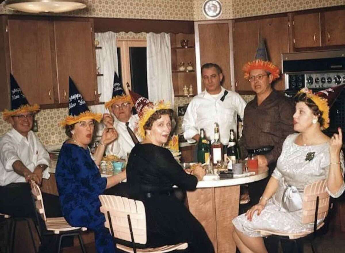 A group of adults in festive hats sit around a kitchen table with drinks and food, celebrating together. The kitchen has retro decor and everyone appears to be enjoying a party.