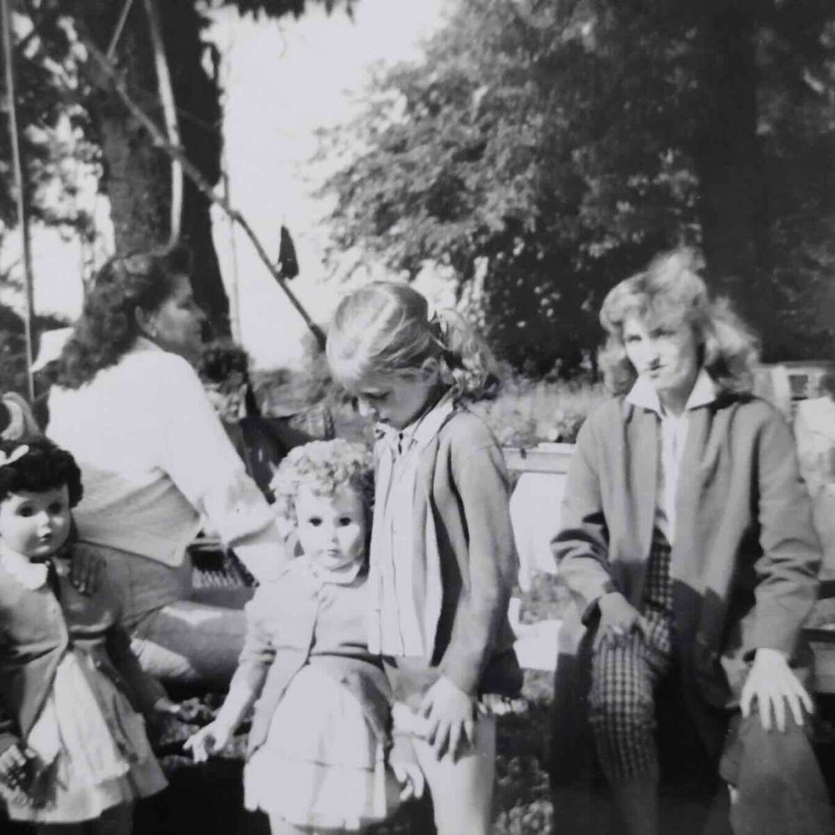 Black-and-white photo of four young girls and an adult outdoors. Three girls stand together in dresses, one with curly hair. Another girl sits on a bench near an adult, with trees and sunlight in the background.