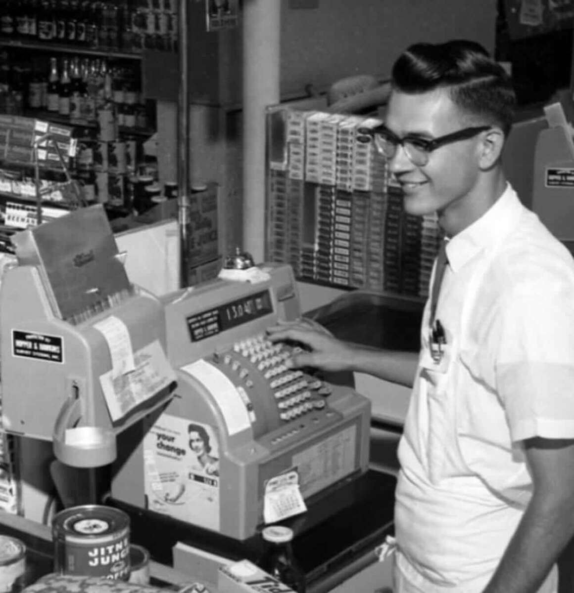 A young man wearing glasses and a white shirt stands smiling behind an old-fashioned cash register in a grocery store, with canned goods and shelves of products in the background.