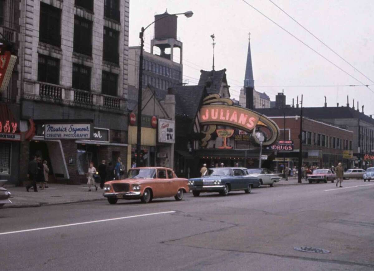 A city street scene from the past with vintage cars parked along the curb, people walking on sidewalks, and storefronts including “Julians” with a large neon sign and a creative photography shop.