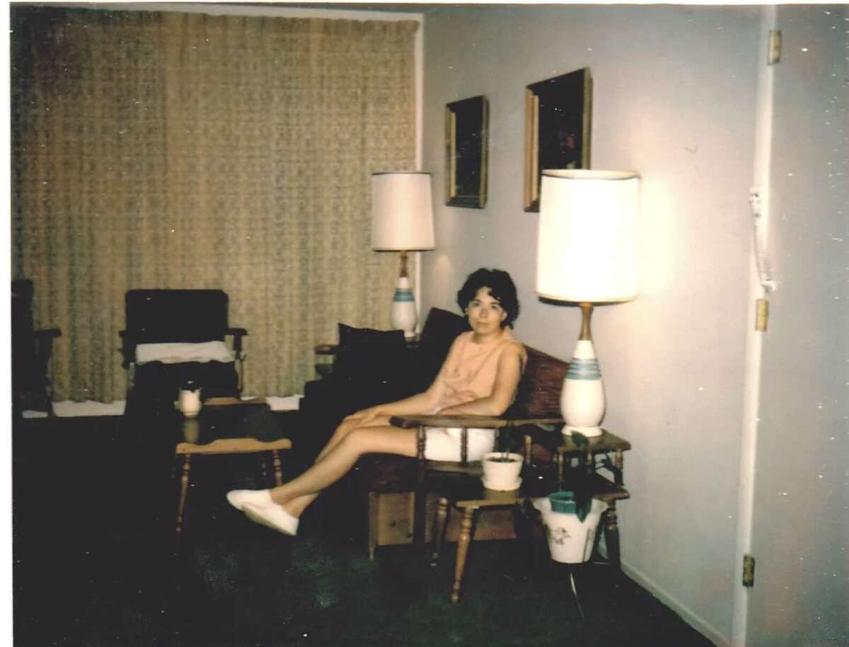 A young woman in a light dress sits on a couch in a living room with two tall lamps, framed pictures on the wall, a wooden coffee table, and a large window with drapes in the background.