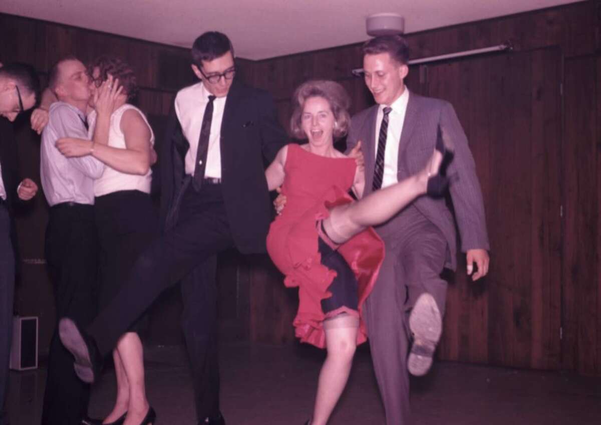 A group of people in formal attire dance energetically indoors; a woman in a red dress lifts her leg playfully while others smile and join in. One couple kisses in the background.