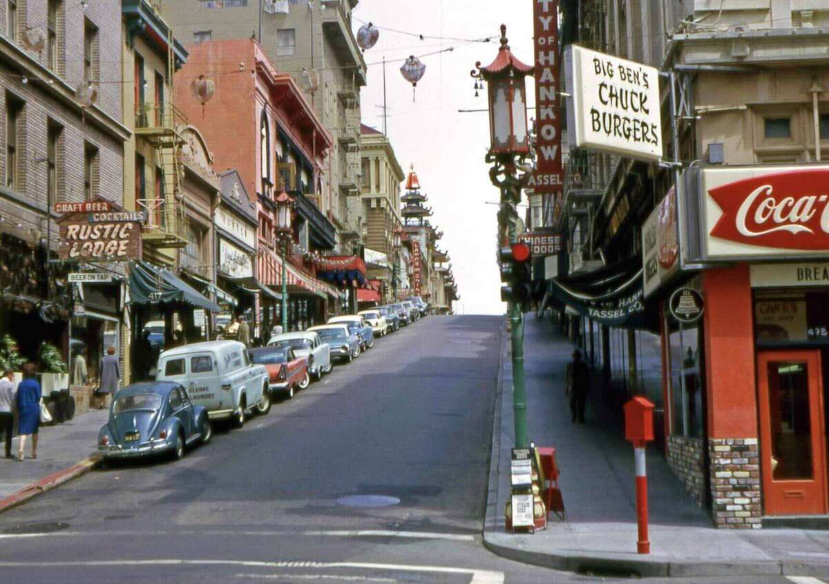 A vintage street scene shows a steep hill lined with classic cars, old storefronts, neon signs, and a Coca-Cola advertisement, capturing a lively city atmosphere reminiscent of mid-20th century Chinatown.