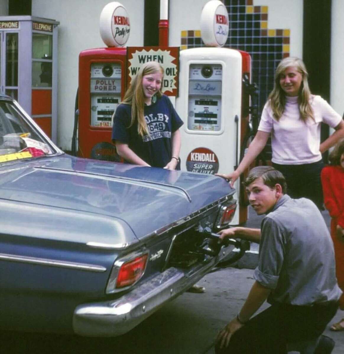 Three young people are at an old-fashioned gas station; two women stand by the open trunk of a blue vintage car while a man crouches, filling the gas tank. Two retro gas pumps are in the background.