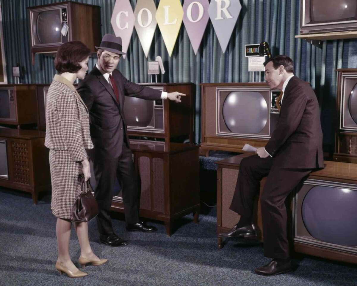 A man in a suit points at a vintage television while speaking to a woman and another man sitting on a wooden TV stand in a 1960s showroom with a “COLOR” banner above several boxy televisions.