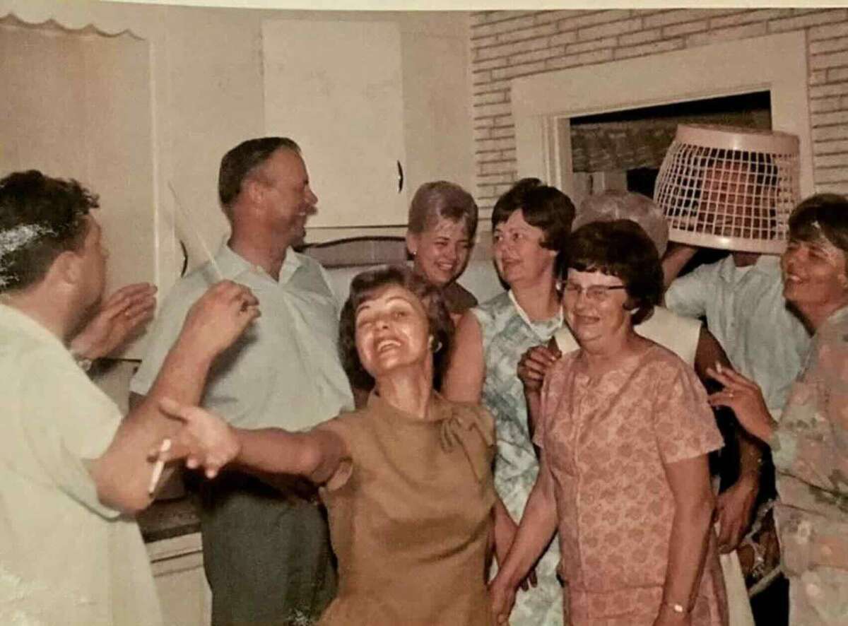A group of people stand smiling and laughing in a kitchen. One person in the back is wearing a laundry basket on their head, adding a playful and humorous touch to the casual gathering.
