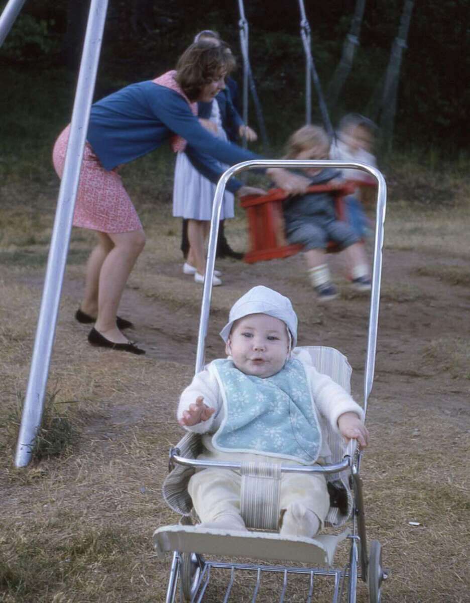 A baby in a stroller sits in the foreground, wearing a blue bib and hat. In the background, a woman pushes two children on swings in a park setting with grass and trees.