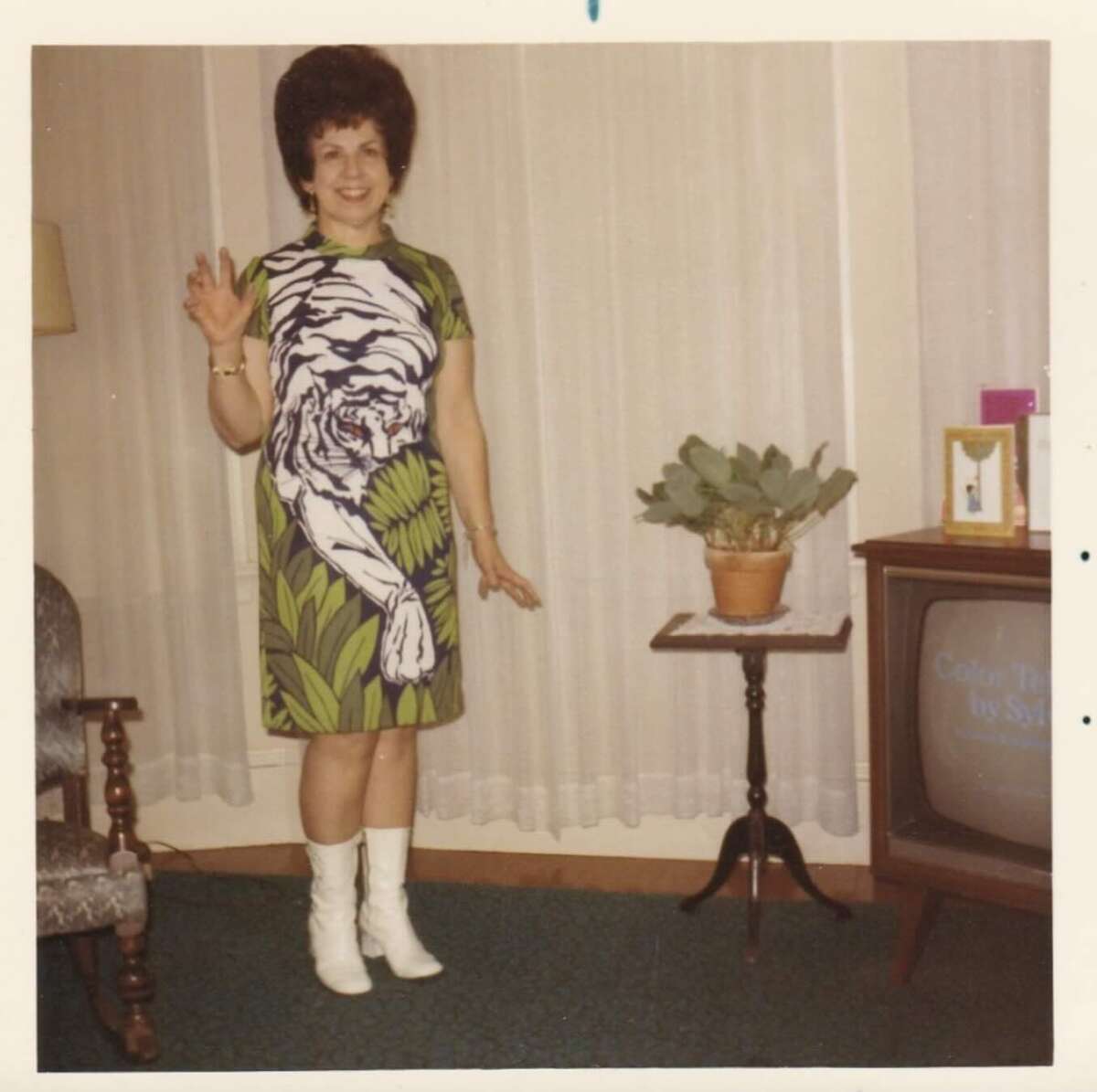 A woman in a green dress with a white tiger design poses indoors, smiling and raising one hand in a playful claw gesture. She wears white boots. A potted plant sits on a small table next to her, near a vintage TV and framed photo.