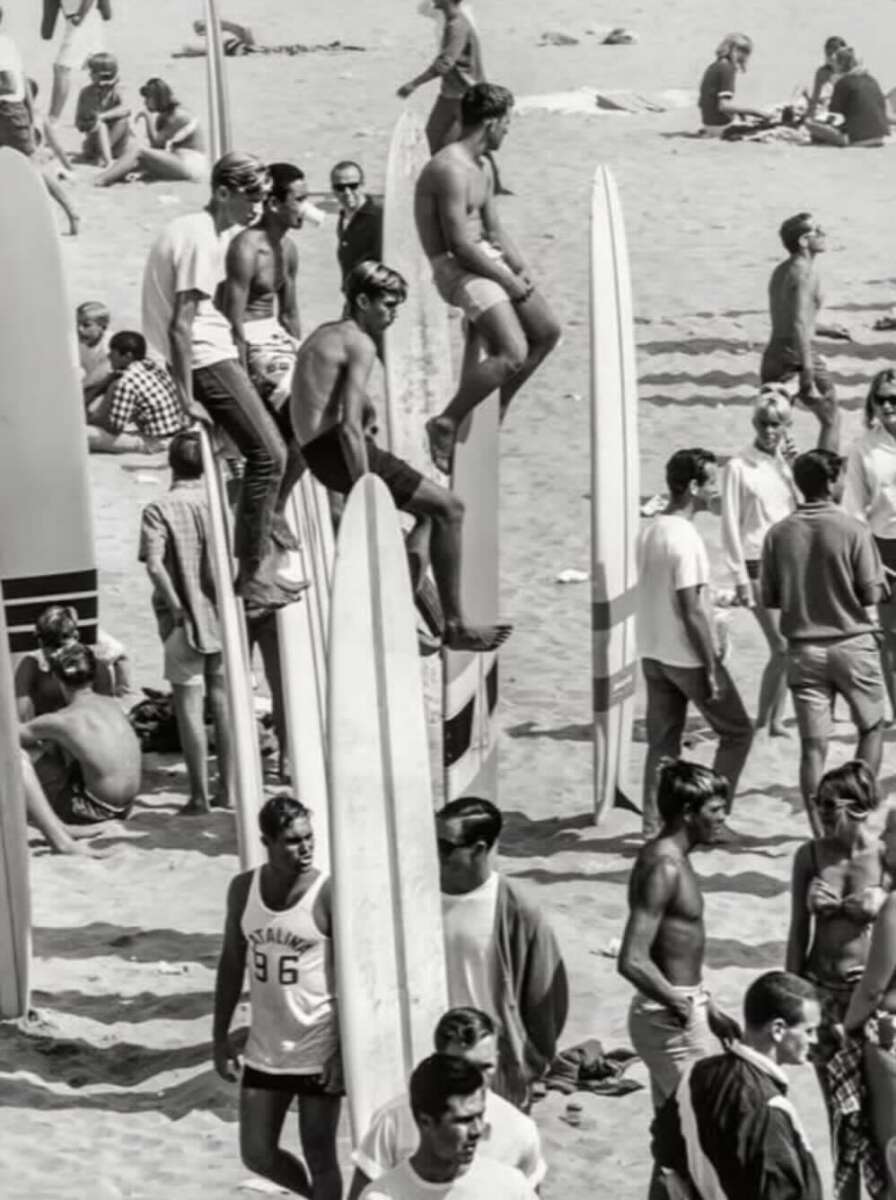 Black and white photo of a crowded beach scene with people standing, walking, and sitting. Several men are perched on upright surfboards, while others gather around, creating a lively and busy atmosphere.