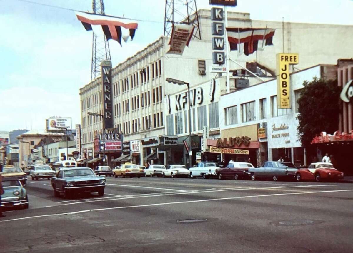 A busy city street from the 1960s with vintage cars, shops, and large signs, including “KFWB,” “Warner,” and “Free Jobs.” Red, white, and black banners hang across the buildings.