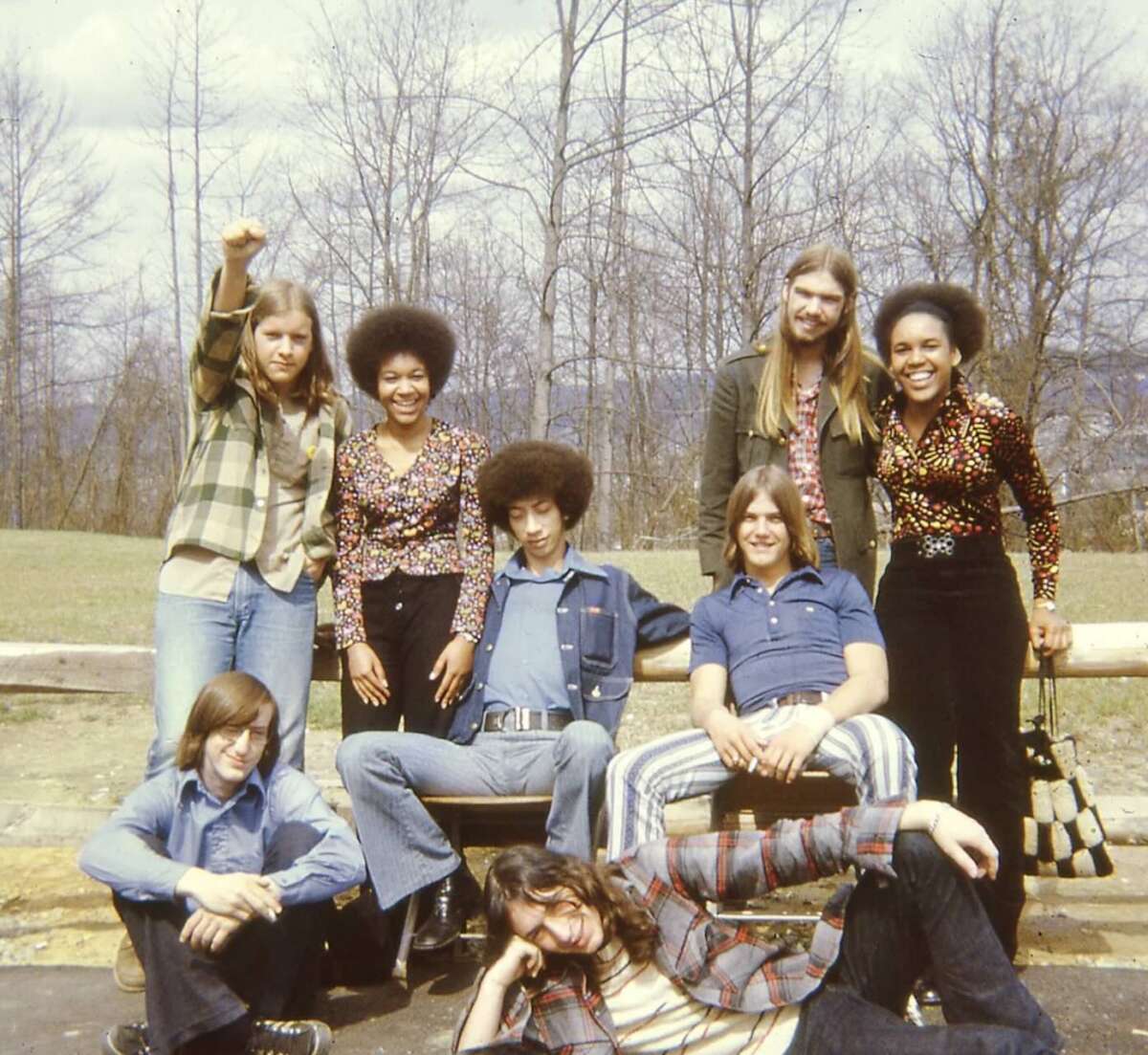 A group of eight young adults pose outdoors in the 1970s; some stand, some sit, and one person raises a fist. They wear colorful, patterned clothes and flared pants, with bare trees and a grassy area in the background.