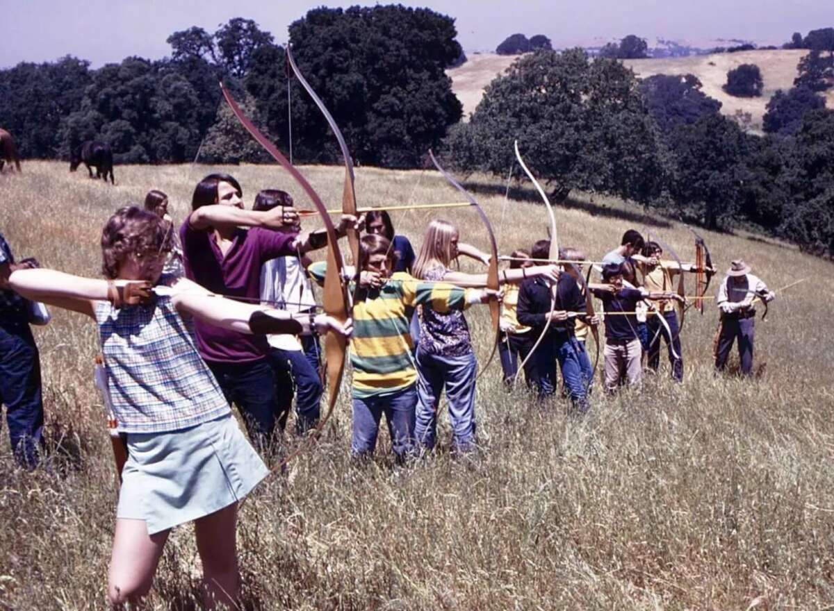 A group of children and teens stand in a line outdoors on a grassy hillside, aiming bows and arrows, with trees and hills in the background. An adult supervises at the end of the line.