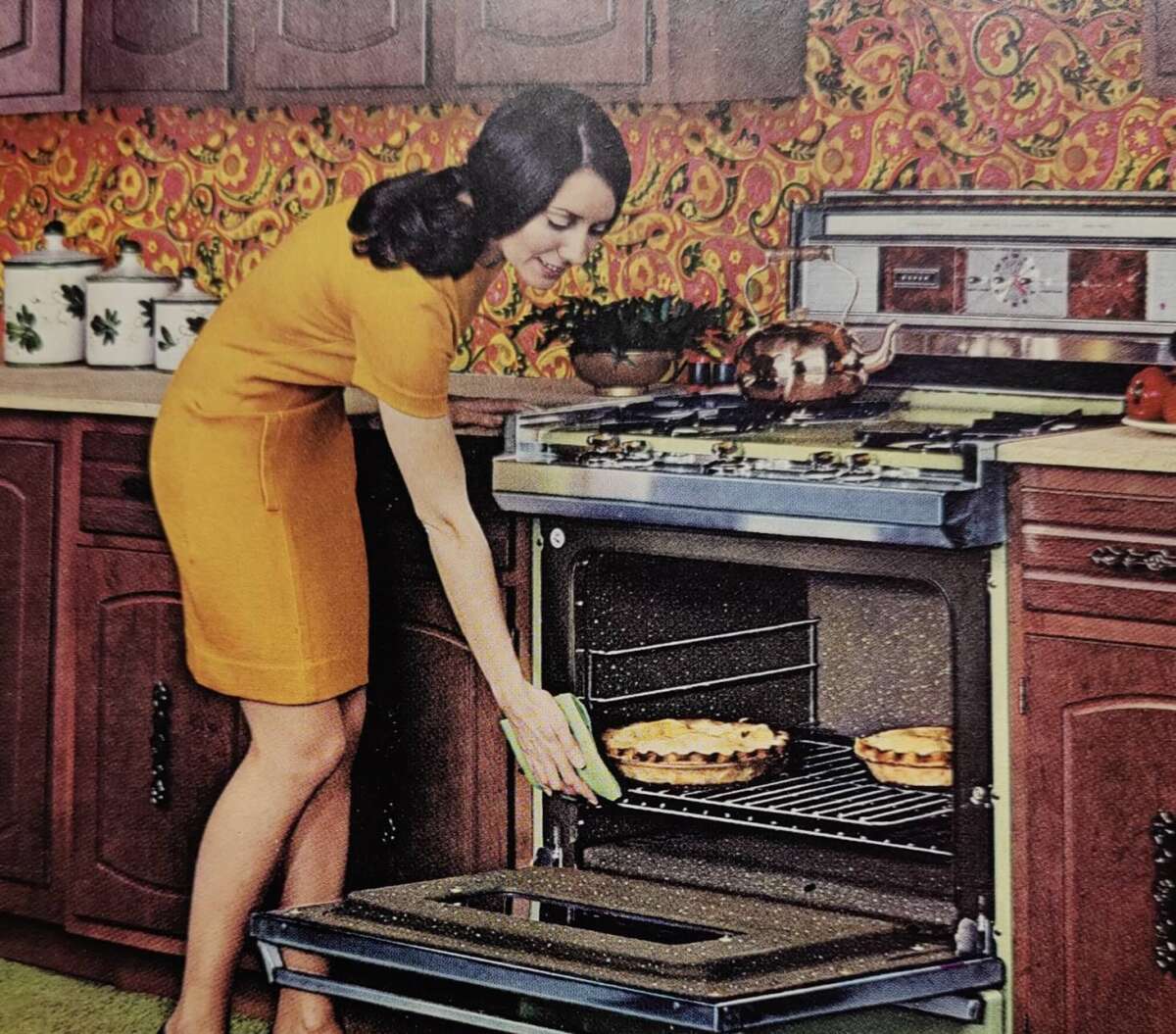 A woman in a yellow dress smiles as she places or removes a pie from an open oven in a retro-style kitchen with wood cabinets and colorful patterned wallpaper.