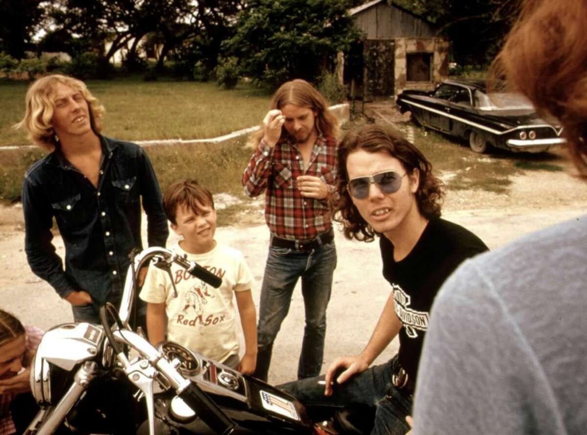 Four men and a boy stand outdoors near a motorcycle. One man wears sunglasses and sits on the bike. The others stand nearby, with trees, grass, and an old car in the background.