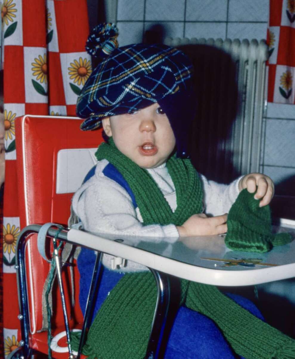 A young child sits in a red high chair, wearing a large plaid cap, a green scarf, and holding a matching green mitten. The background features red curtains with yellow flowers and a white tiled wall.
