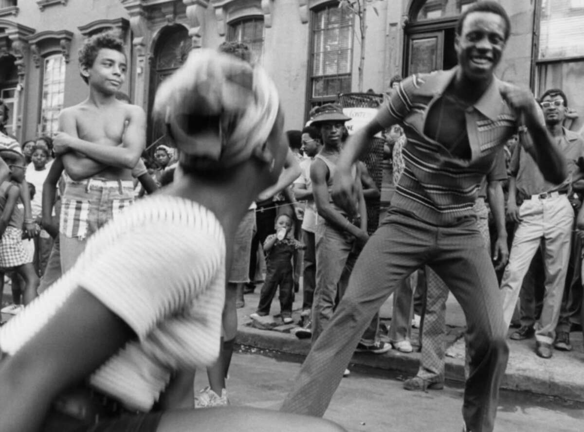 Black-and-white photo of a lively street scene where a man and woman dance joyfully, surrounded by smiling and watching people on a city sidewalk in front of apartment buildings.