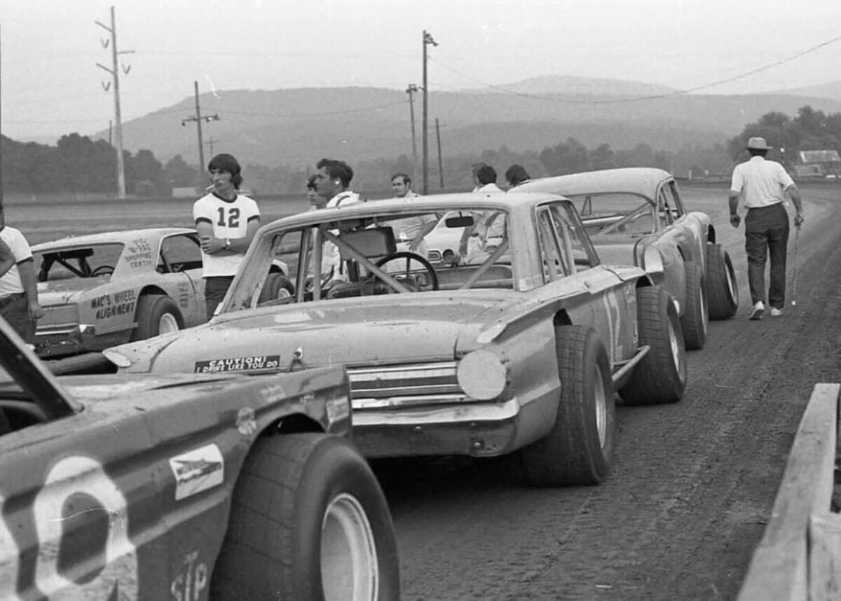 Black and white photo of several vintage stock cars lined up on a dirt racetrack, with drivers and crew members standing nearby. Trees, utility poles, and hills are visible in the background.
