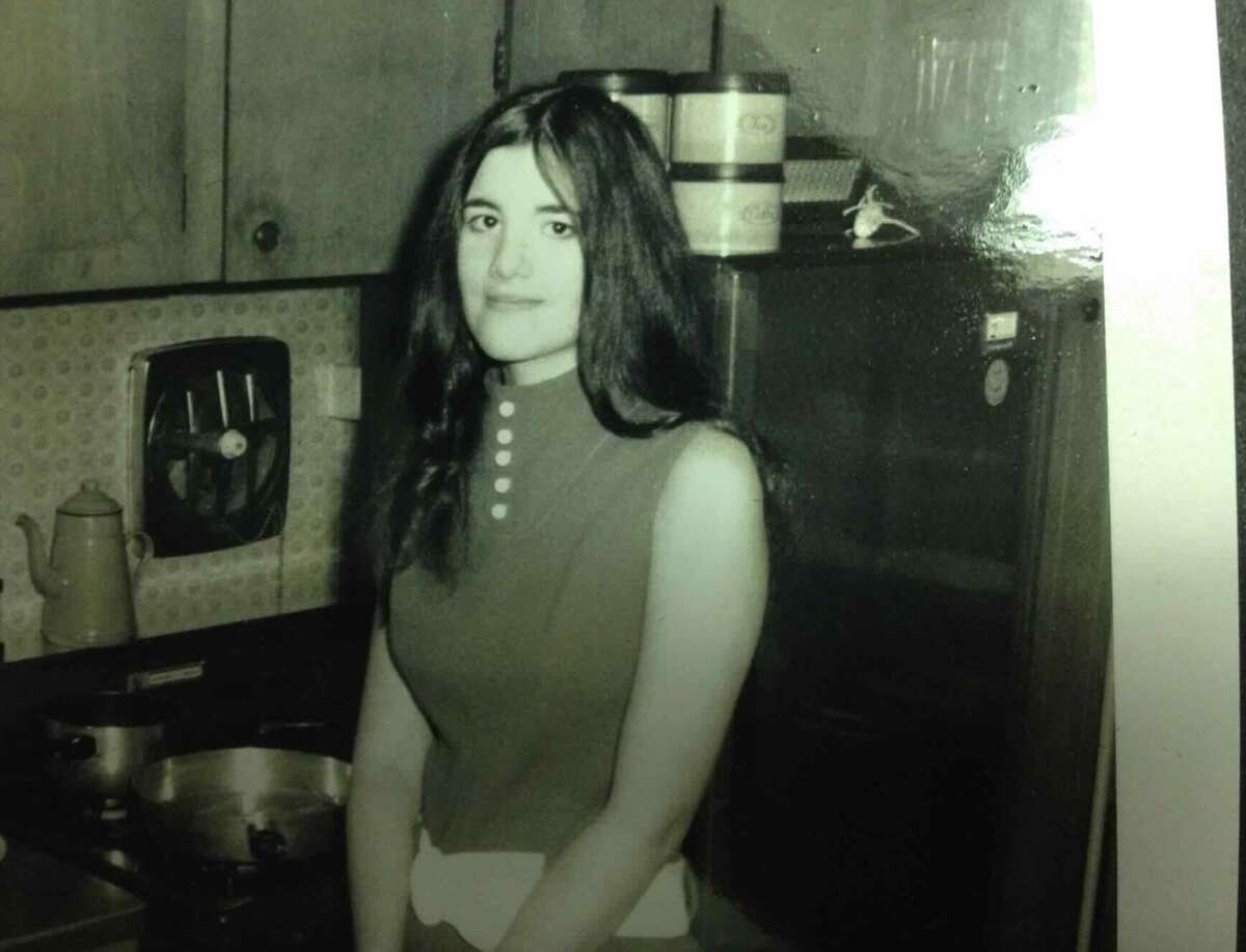 A young woman with long dark hair stands in a vintage kitchen, wearing a sleeveless top. Behind her are a refrigerator, a pot on the stove, and shelves with canisters. The photo is black and white.