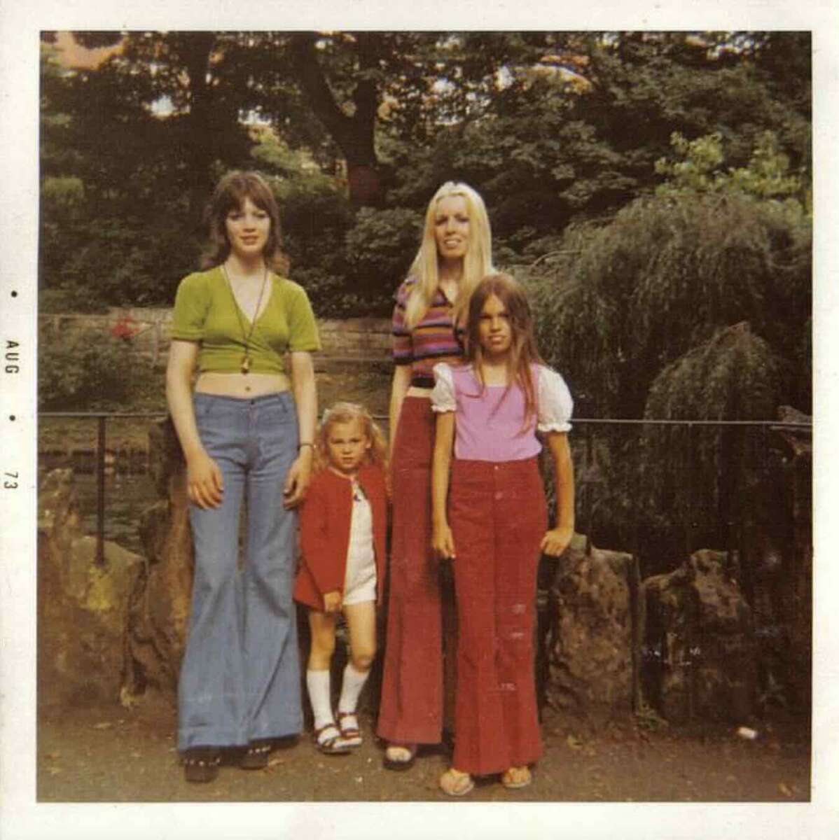 Four people stand together outdoors near rocks and trees, wearing colorful 1970s clothing. The photo, dated August 1973, shows three girls and one woman posing for the camera in a green, park-like setting.