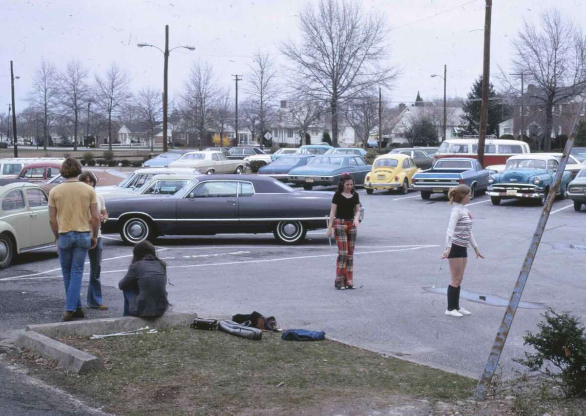A group of teens hang out in a 1970s parking lot, with several vintage cars parked nearby. Two girls play with a jump rope while others stand and sit nearby. Leafless trees and houses are in the background.
