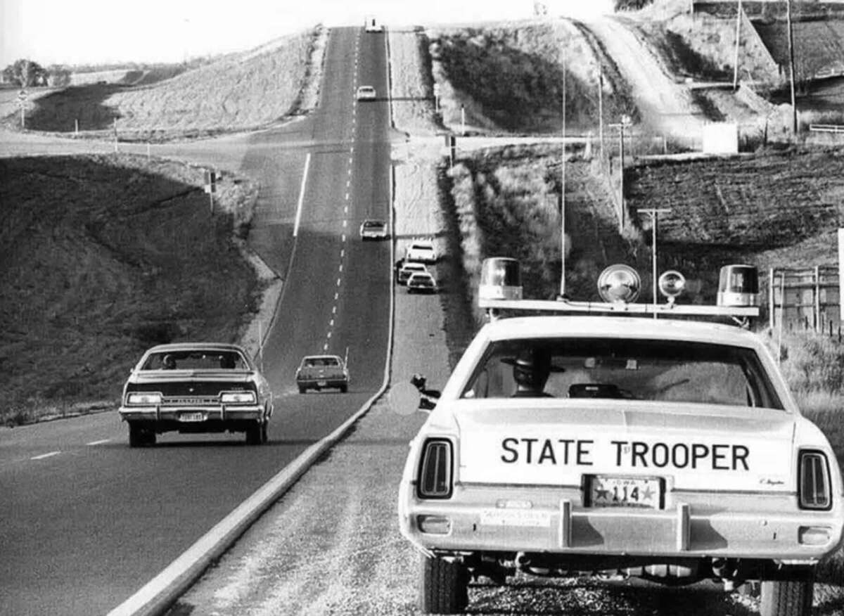 A vintage black-and-white photo shows a "State Trooper" car parked on the shoulder of a rural highway, with other cars driving along the road and rolling hills in the background.